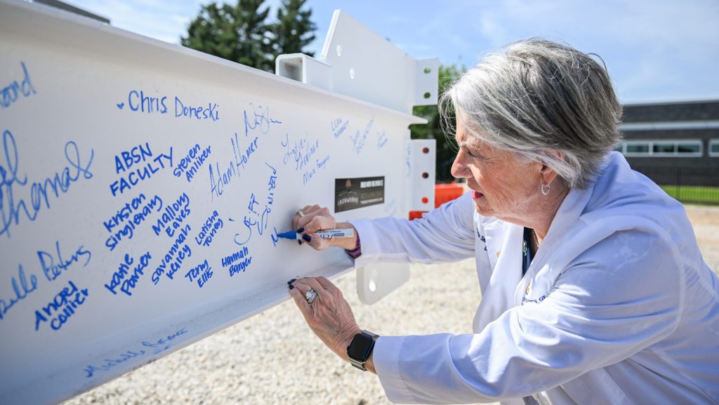 An individual in a white coat signs a large white beam outdoors, writing names in blue marker.